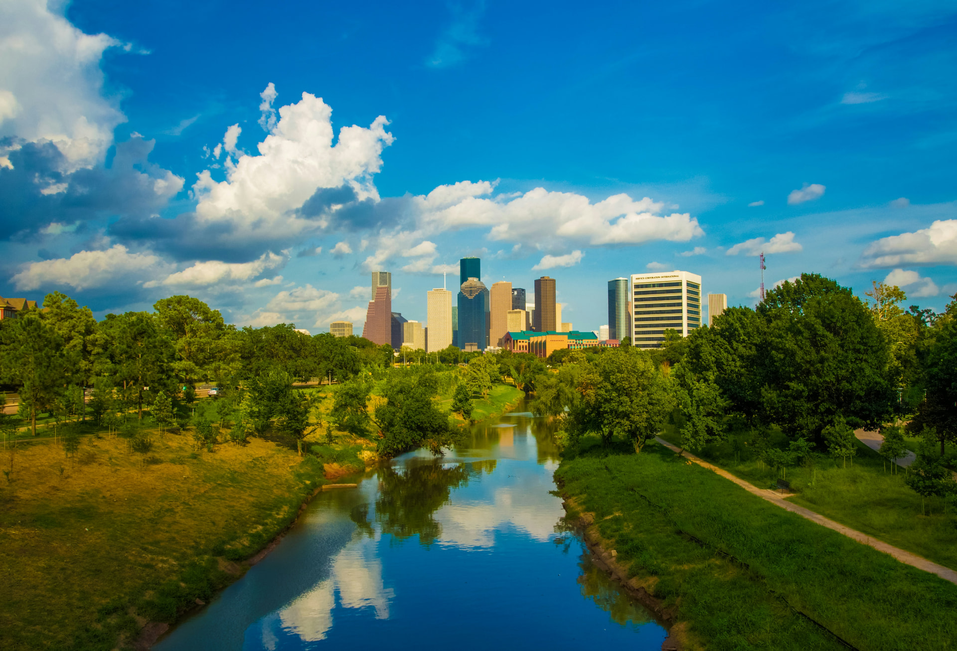Houston skyline over a tree-lined bayou under a bright blue sky